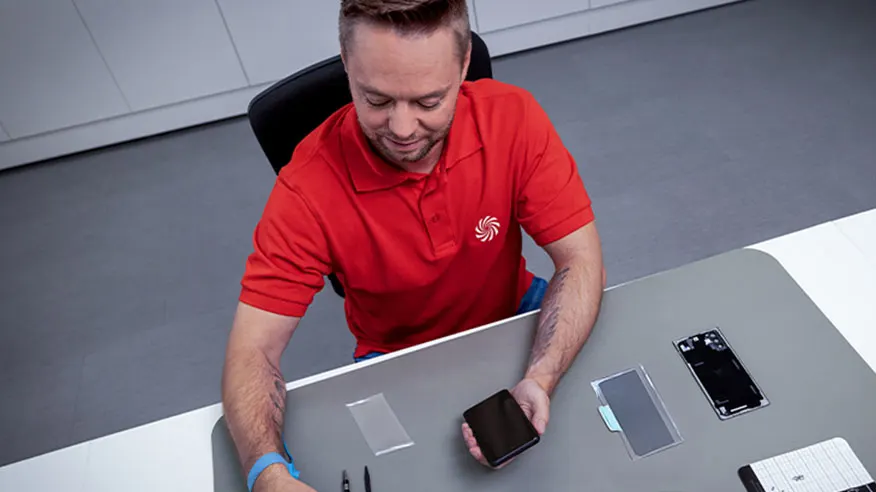 Mężczyzna w czerwonej koszulce polo siedzi przy stole z telefonami i akcesoriami do ich naprawy na szarym podkładzie.