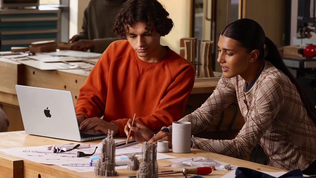 Młodzi mężczyzna i kobieta przed MacBookiem Pro 2025 ustawionym na biurku z projektami w formie wydruków na papierze i 3D.