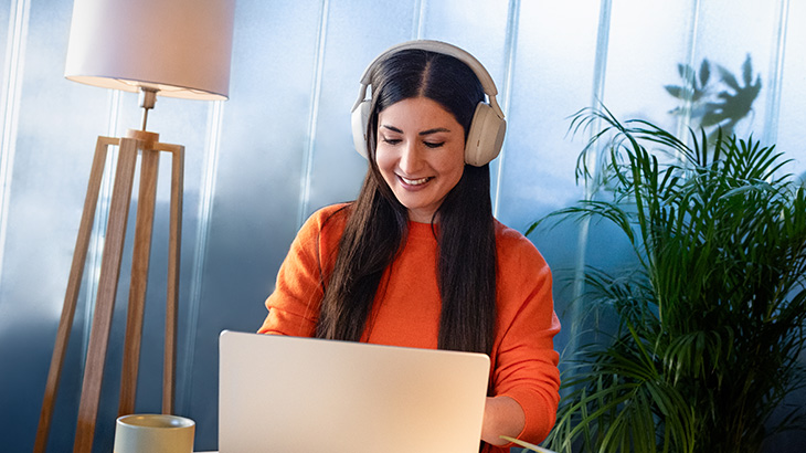 Kobieta z ciemnymi długimi włosami, z nausznymi słuchawkami siedzi uśmiechnięta przed laptopem. W tle rośliny i lampa