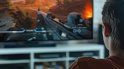 Chłopiec ogląda na telewizorze grę akcji. Telewizor stoi na białej szafce RTV. W tle widać okno i kanapę.