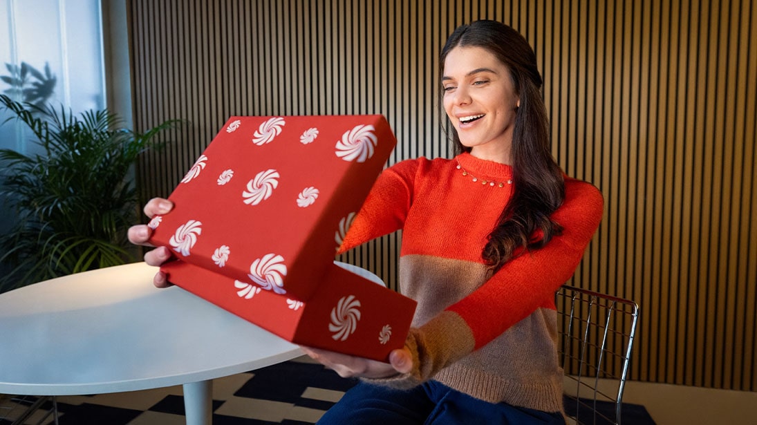 Uśmiechnięta kobieta otwiera czerwone pudełko z białymi wzorami na tle ściany z drewnianych paneli i zielonej rośliny.