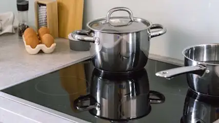 Dwa stalowe garnki na czarnej płycie indukcyjnej. Na blacie leżą jajka w pojemniku i biała ścierka.