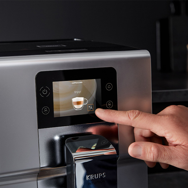 Srebrny ekspres do kawy z czarnym panelem dotykowym. Palec naciska ikonę filiżanki cappuccino na ekranie.