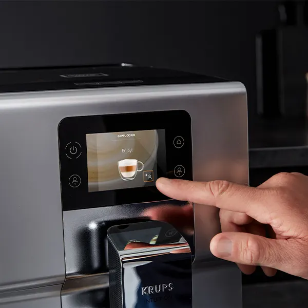 Srebrny ekspres do kawy z czarnym panelem dotykowym. Palec naciska ikonę filiżanki cappuccino na ekranie.