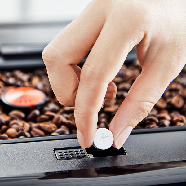 Dłoń wkłada biały okrągły przedmiot do czarnego urządzenia; w tle brązowe ziarna kawy i pomarańczowa kapsułka.