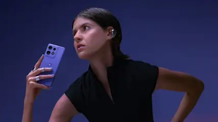 Kobieta trzyma fioletowy telefon z trzema obiektywami. Ma czarną bluzkę i srebrne pierścionki. Tło jest fioletowe.