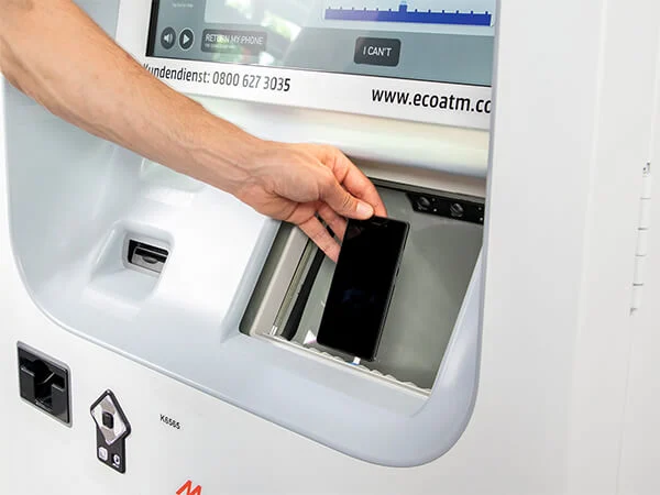 Osoba wkłada czarny telefon do białego automatu ecoATM z ekranem i przyciskami. Automat ma logo i numer infolinii.