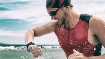 Mężczyzna w czerwonym stroju triathlonowym i czepku pływackim wchodzi do wody, na nadgarstku ma czarny zegarek. W tle morze i góry.