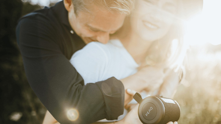 Mężczyzna w czarnej koszuli obejmuje kobietę w białej bluzce. W dłoni trzyma czarny głośnik przenośny na tle zieleni.