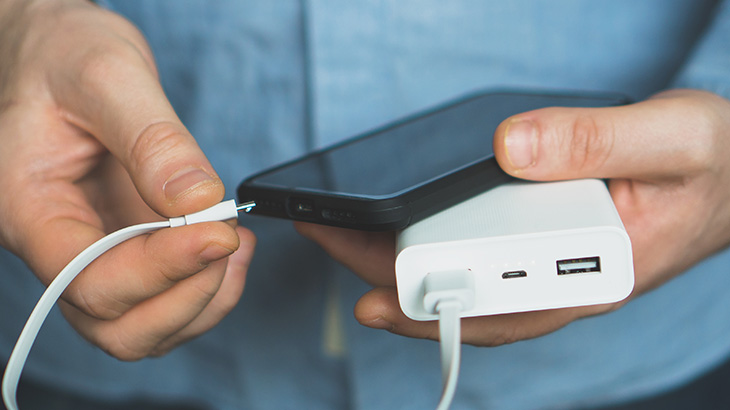 Zbliżenie na dłonie podłączające czarny smartfon za pomocą białego kabla do przenośnego, białego powerbanku.