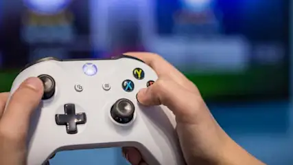 Dłonie trzymają biały kontroler Xbox. W tle widać nieostry ekran telewizora na tle niebieskiej ściany.
