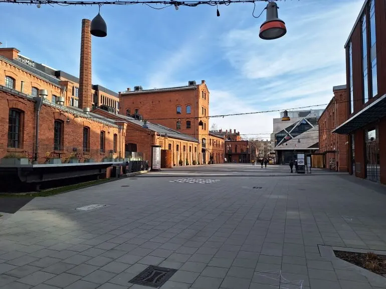 Ulica z ceglanymi budynkami po bokach i brukowanym placem. W tle widoczna nowoczesna architektura i niebo.