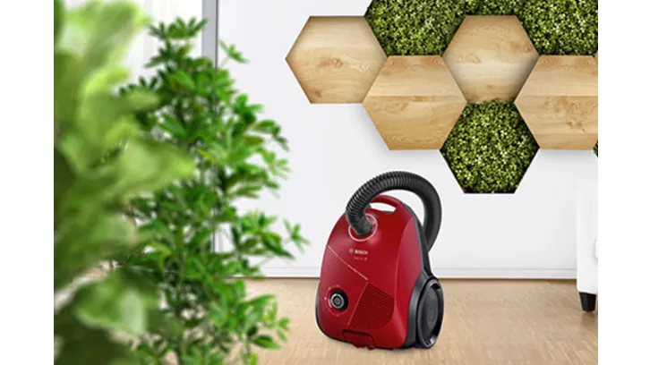 Czerwony odkurzacz obok zielonej rośliny. Na ścianie dekoracja z drewnianych i zielonych sześciokątów.