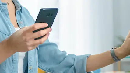 Osoba w dżinsowej koszuli trzyma telefon obok pralki z okrągłymi drzwiami w jasnym wnętrzu.