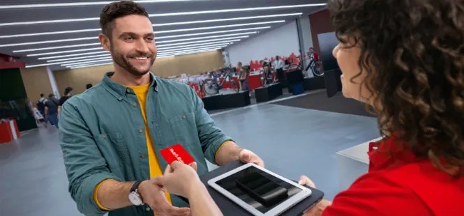 Mężczyzna wręcza kartę i tablet osobie w czerwonej koszuli, w tle widoczni ludzie i oświetlenie sufitowe.