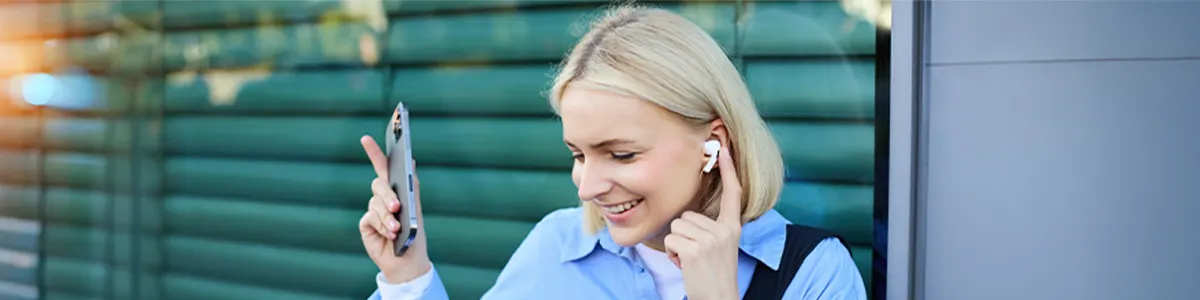 Kobieta w niebieskiej koszuli trzyma telefon. Uśmiecha się, ma słuchawkę w uchu. W tle zielona ściana.