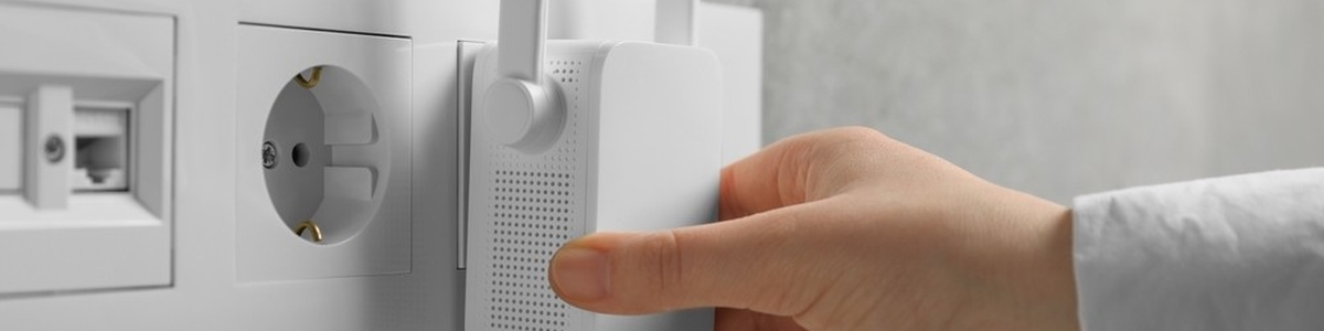 Biały wzmacniacz WiFi w gniazdku elektrycznym, obok gniazdo telefoniczne. Ręka wkłada urządzenie do gniazdka. Tło szare.