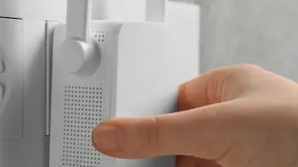 Biały wzmacniacz WiFi w gniazdku elektrycznym, obok gniazdo telefoniczne. Ręka wkłada urządzenie do gniazdka. Tło szare.