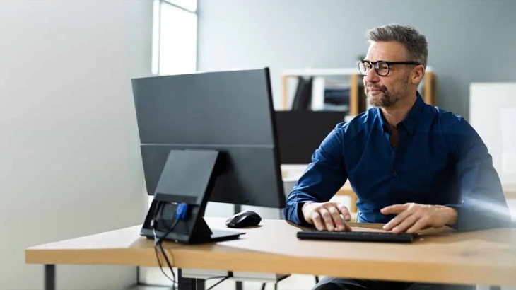 Mężczyzna w okularach używa komputera stacjonarnego z klawiaturą i myszą na drewnianym biurku w biurze.