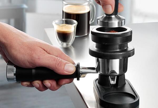 Przygotowanie kawy: ręka trzyma uchwyt, ubijak nad kolbą, w tle filiżanka espresso i szklanka kawy.