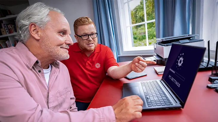 Dwaj mężczyźni przy biurku patrzą na laptopa. Jeden wskazuje na ekran. W tle okno, drukarka i router.