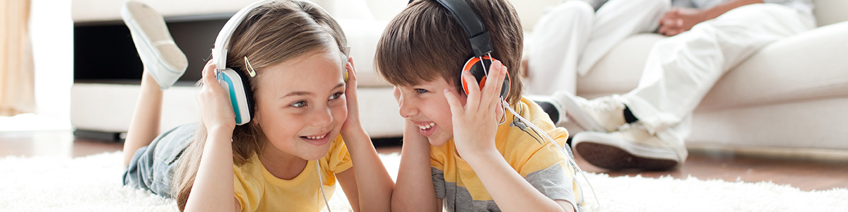 Dzieci leżą na dywanie w słuchawkach, słuchając muzyki, za nimi siedzi osoba na białej kanapie.