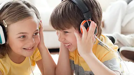 Dzieci leżą na dywanie w słuchawkach, słuchając muzyki, za nimi siedzi osoba na białej kanapie.