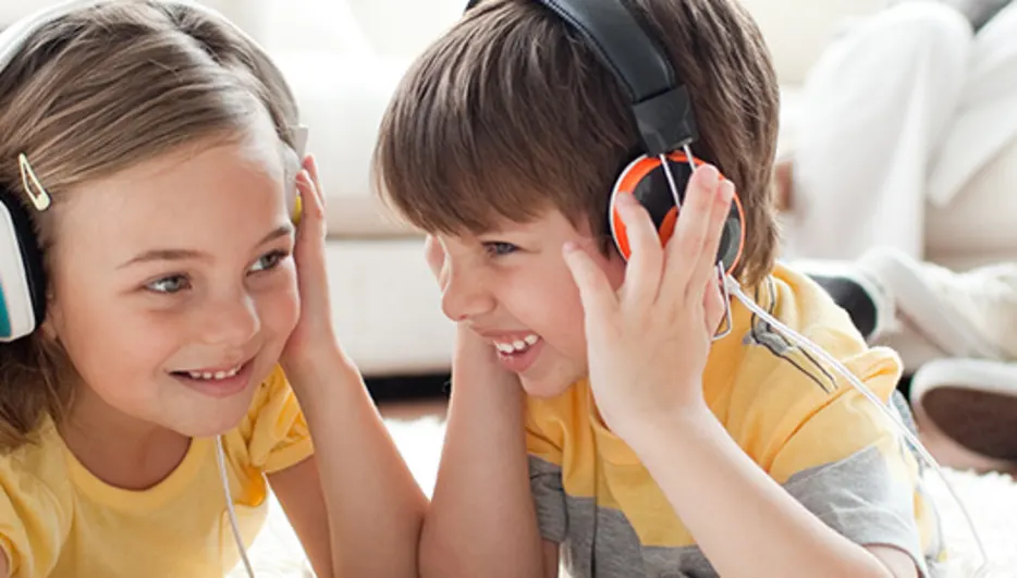 Dzieci leżą na dywanie w słuchawkach, słuchając muzyki, za nimi siedzi osoba na białej kanapie.