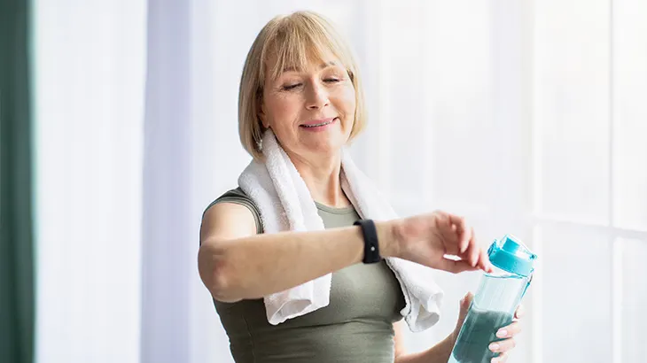 Kobieta z ręcznikiem na szyi, sprawdza zegarek. Trzyma butelkę z wodą na tle okna.