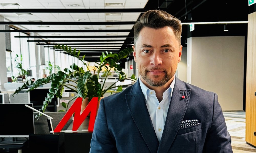 Mężczyzna w garniturze na tle biura z roślinami i czerwonym logo. W tle biurka i okna.