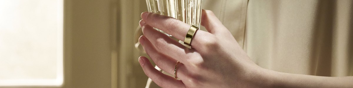 Dłoń trzyma szklanki, na palcach złote pierścionki, tło beżowe.