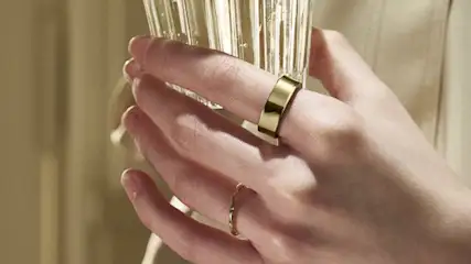 Dłoń trzyma szklanki, na palcach złote pierścionki, tło beżowe.