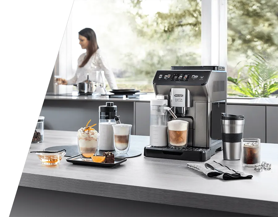 W kuchni stoi ekspres do kawy, obok szklanki z kawą i akcesoria. W tle kobieta gotuje przy oknie.