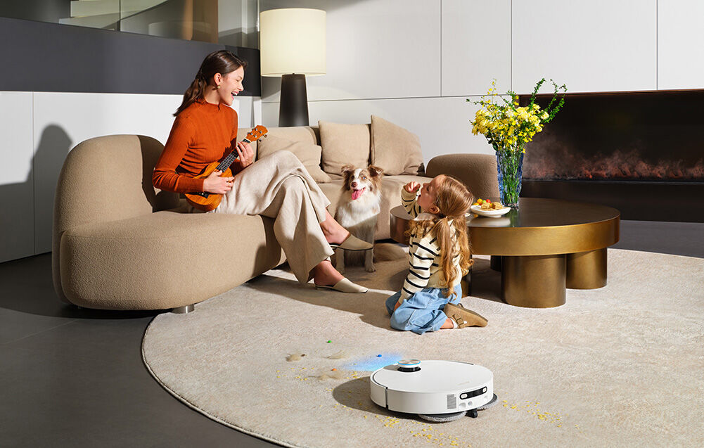 Na grafice widać mamę z córką bawiące się w salonie gdzie obok odkurza robot Dreame.