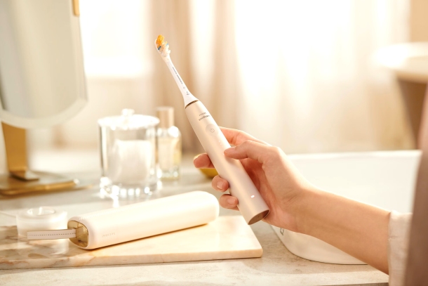 Na blacie leży białe etui obok uchwytu na szczoteczkę. Kobieta trzyma szczoteczkę do zębów. W tle widać szklankę.