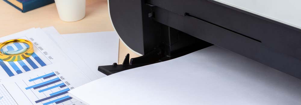 Drukarka drukuje dokument z wykresami słupkowymi; obok leży lupa i kubek.