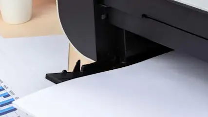 Drukarka drukuje dokument z wykresami słupkowymi; obok leży lupa i kubek.