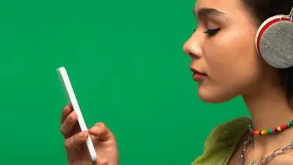 Kobieta w słuchawkach trzyma telefon na zielonym tle. Ma na sobie naszyjniki, w tym kolorowe koraliki.