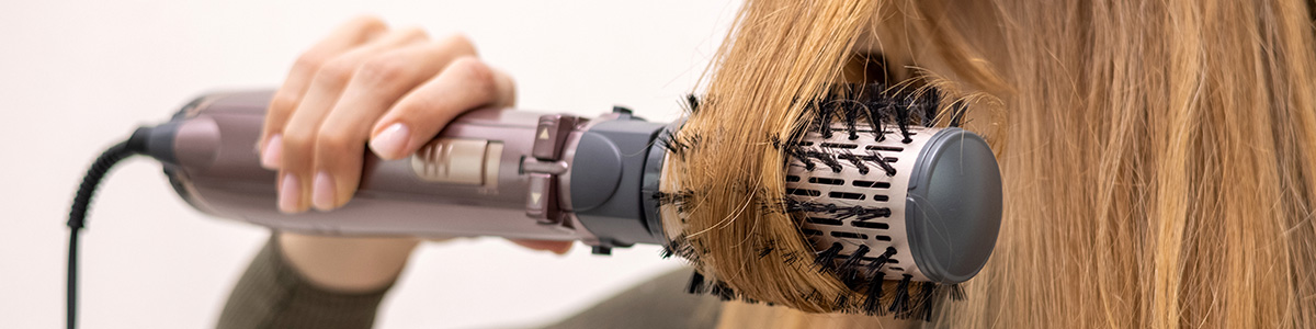 Kobieta używa okrągłej szczotki do modelowania włosów. Włosy są nawinięte na szczotkę, a w dłoni trzymana jest suszarka.
