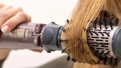 Kobieta używa okrągłej szczotki do modelowania włosów. Włosy są nawinięte na szczotkę, a w dłoni trzymana jest suszarka.