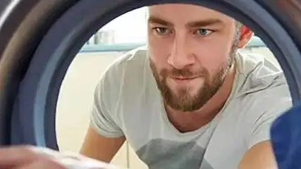 Mężczyzna w białej koszulce patrzy do pralki, w której widać niebieskie ubrania.