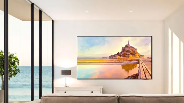 Salon z widokiem na morze; telewizor pokazuje obraz zamku na tle nieba. Obok lampa na komodzie, roślina w doniczce i okno.