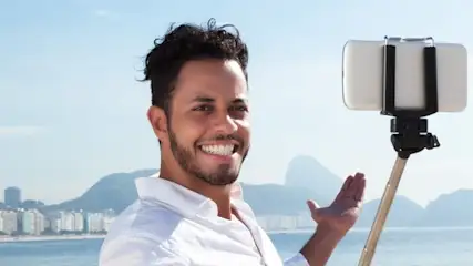 Mężczyzna z kijem do selfie na tle plaży, wody i zabudowań miejskich.
