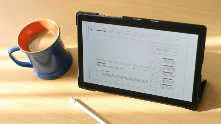 Na biurku stoi niebieski kubek z napojem, tablet z tekstem i biały rysik.