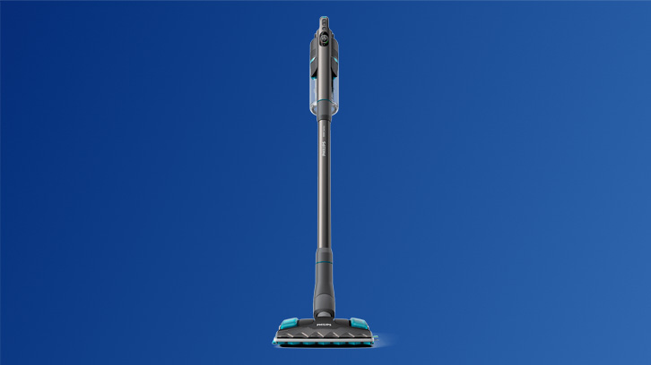 Na niebieskim tle stoi pionowo szary odkurzacz z turkusowymi akcentami na szczotce i uchwycie.