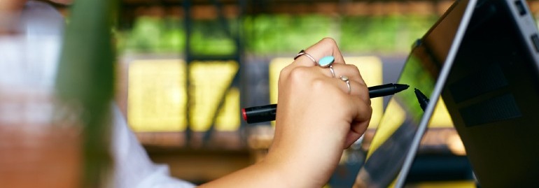 Dłoń z pierścionkami trzyma czarny rysik na ekranie tabletu. W tle zielona roślinność i żółte elementy.