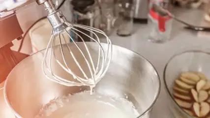 Metalowy mikser z trzepaczką w misce z białym kremem. Obok miska z pokrojonymi jabłkami, słoiki i przyprawy na blacie.