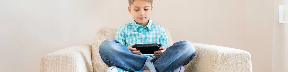 Chłopiec w koszuli w kratę siedzi na fotelu ze skrzyżowanymi nogami, trzymając czarną konsolę do gier.
