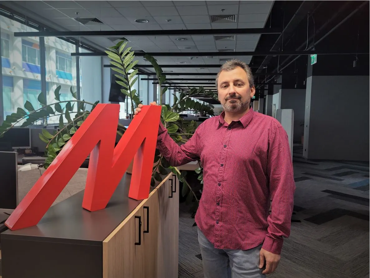 Mężczyzna w koszuli w kratę opiera się o czerwone logo M na szafce w biurze z roślinami i oknami w tle.