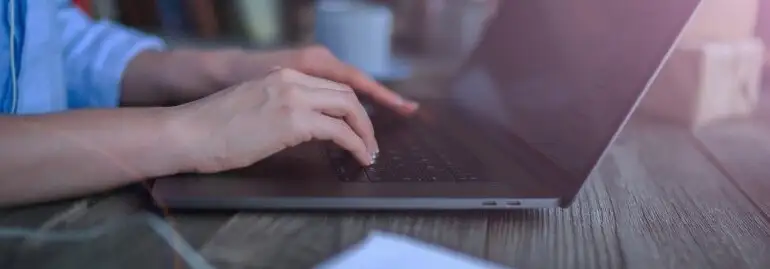 Osoba pisze na laptopie na drewnianym stole. W tle widać książki i białe przedmioty.